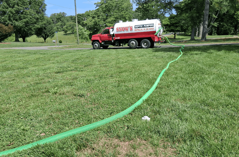 Snow's Septic Pumping red and white vacuum tanker truck parked roadside with the green suction hose run across a lawn to a septic tank